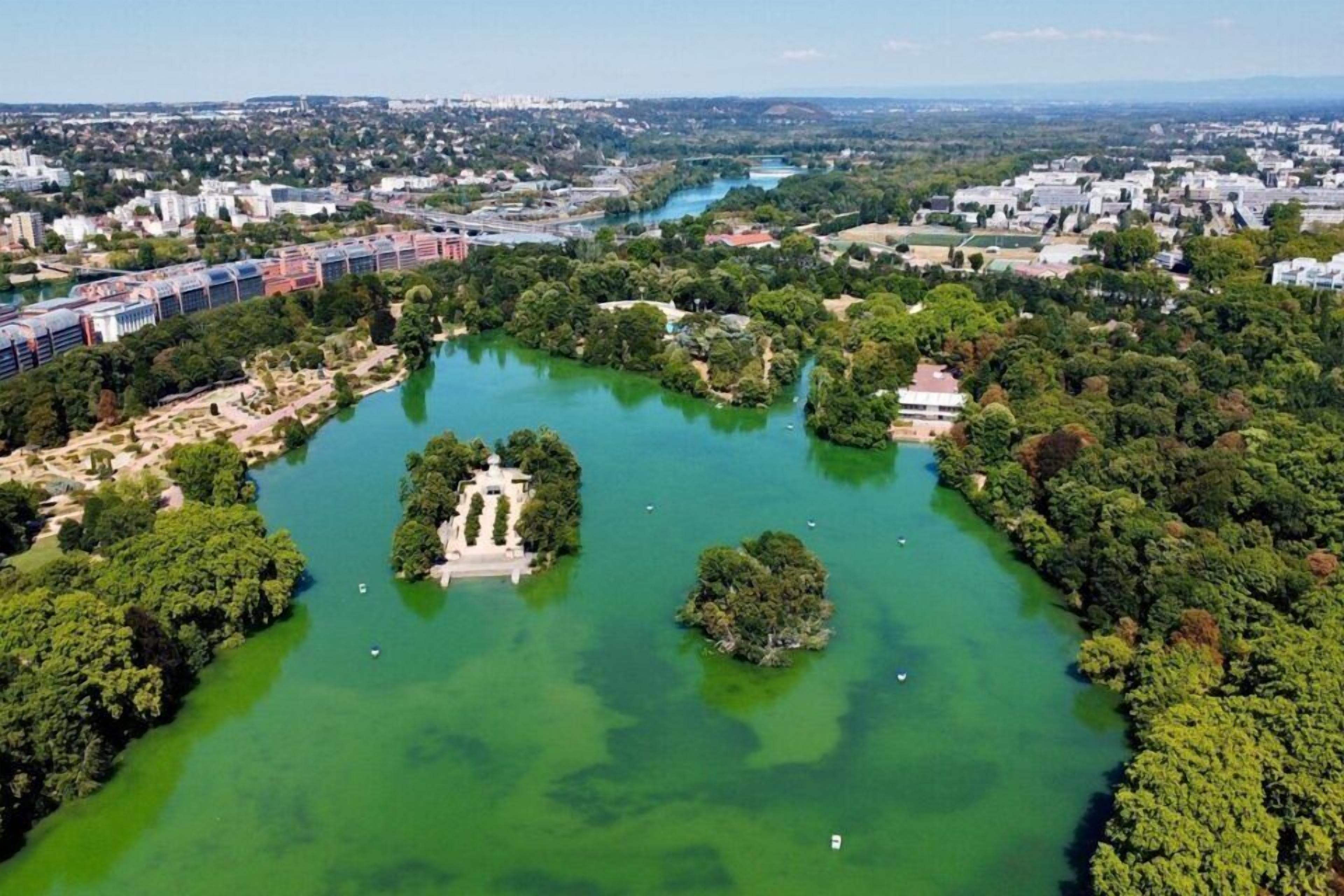 Ein Luftbild eines Parks mit einer zentralen Insel, umgeben von Wasser und Grünflächen, mit Gebäuden im Hintergrund.