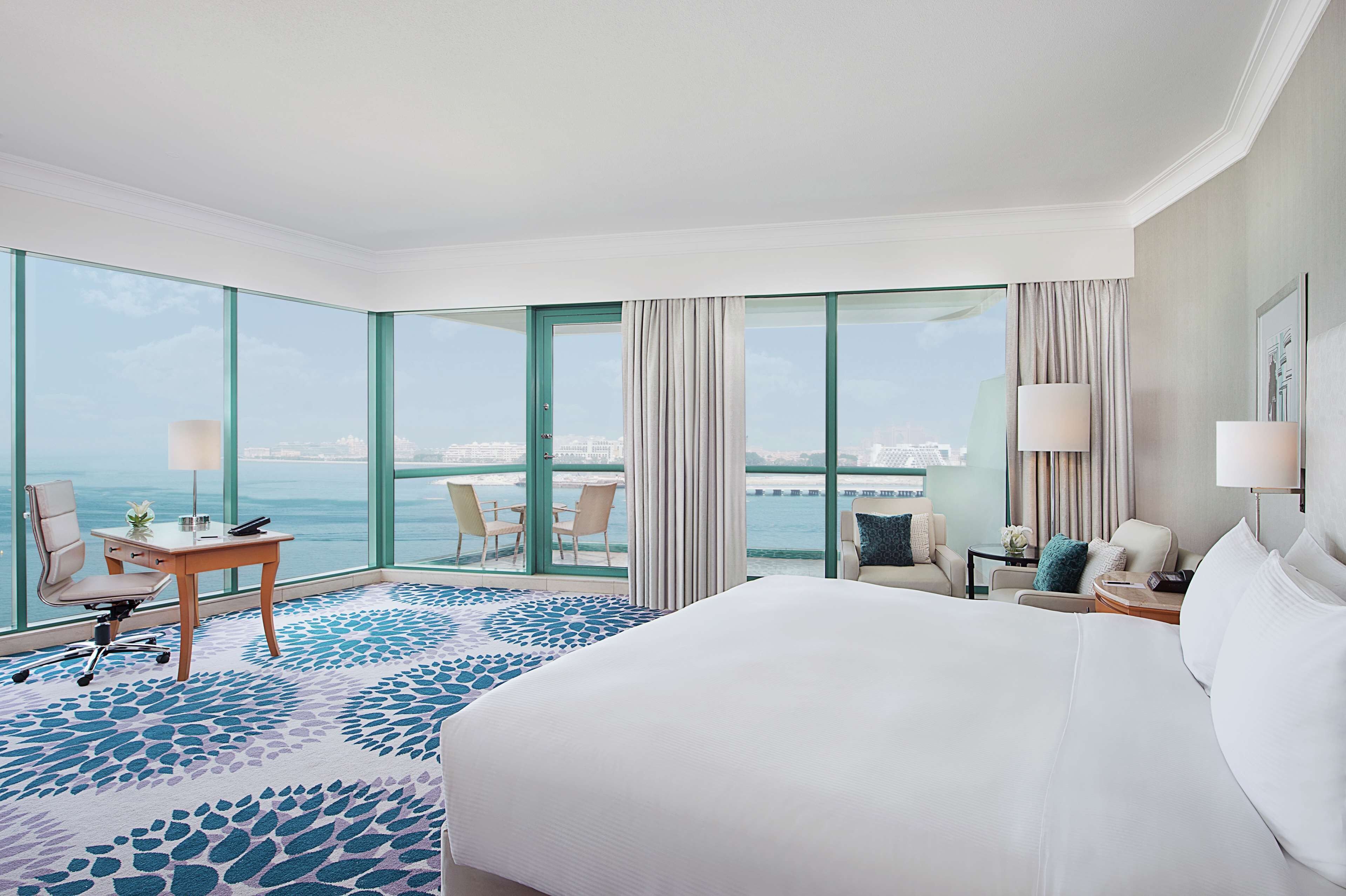 Een hotelkamer met een groot bed, een bureau, een stoel en uitzicht op de zee door grote ramen.