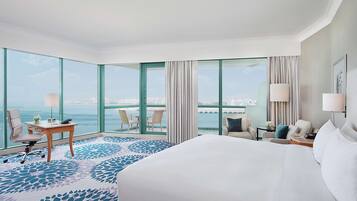 Een hotelkamer met een groot bed, een bureau, een stoel en uitzicht op de zee door grote ramen.