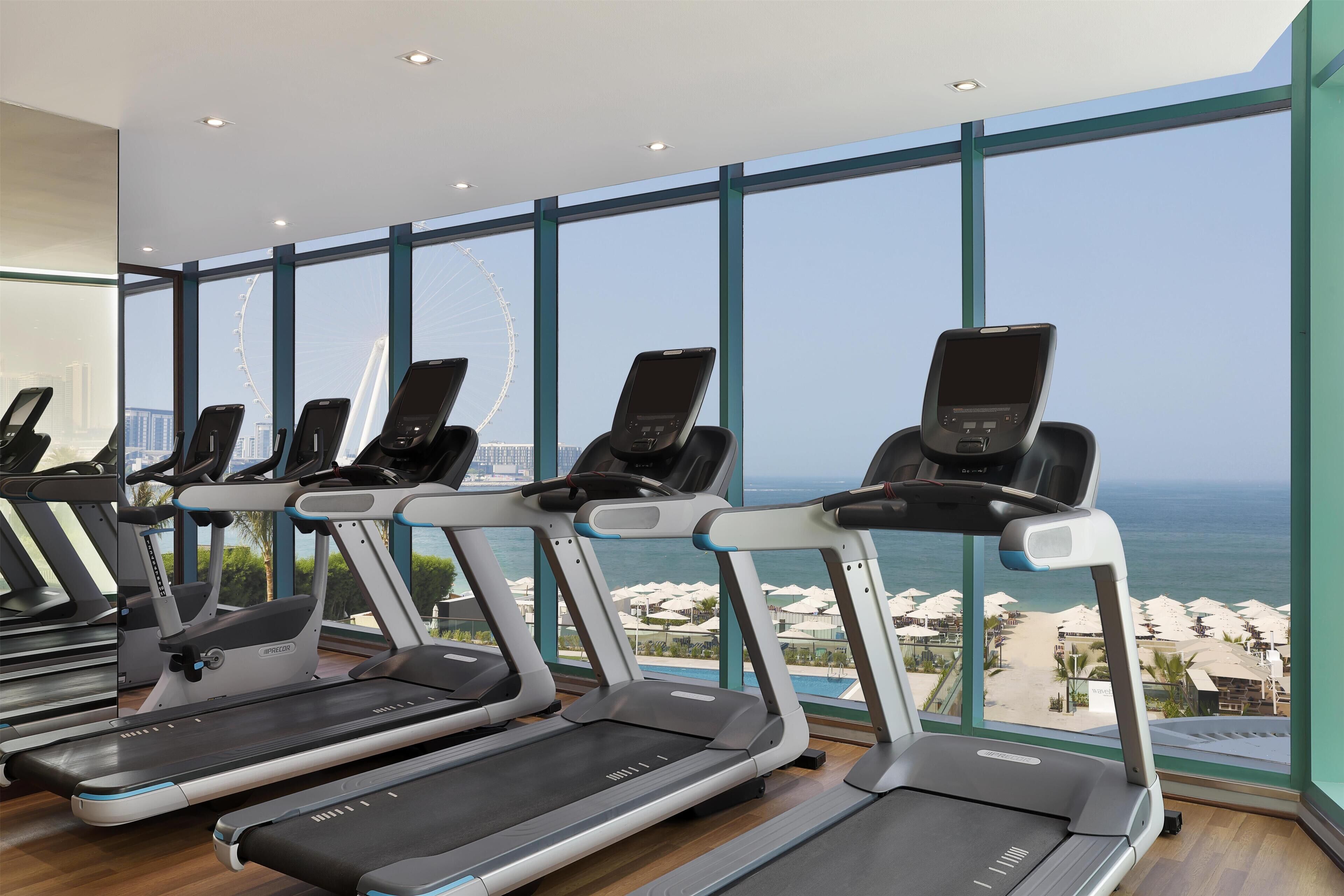 A gym with treadmills and a large window overlooking a beach and a Ferris wheel.