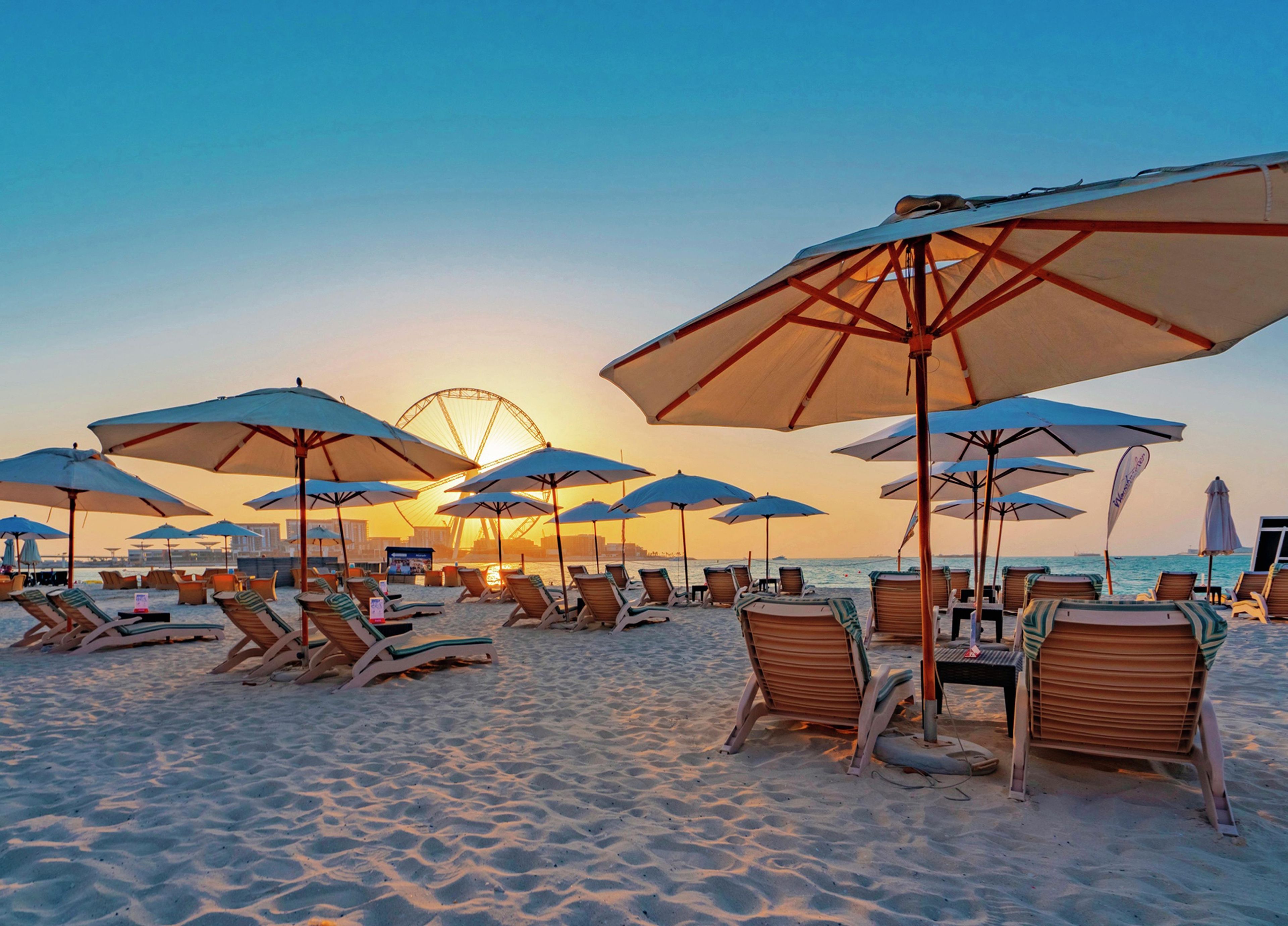 A beach with sun loungers and umbrellas, a Ferris wheel in the background, and a clear sky.