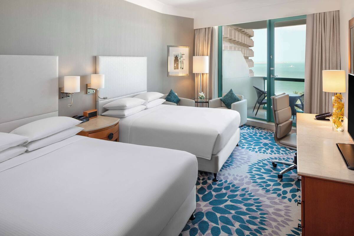 A hotel room with two beds, a desk, a chair, and a view of the sea.
