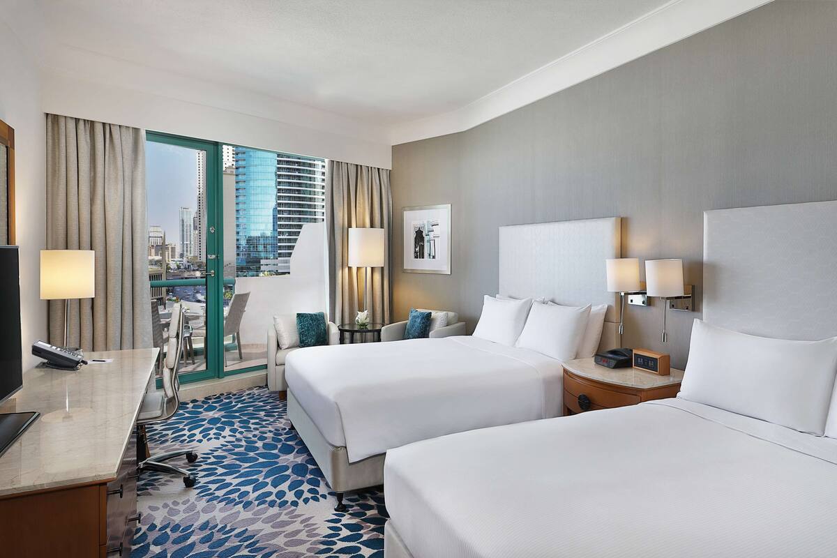 A hotel room with two beds, a desk with a computer, a chair, a lamp, and a view of a cityscape.