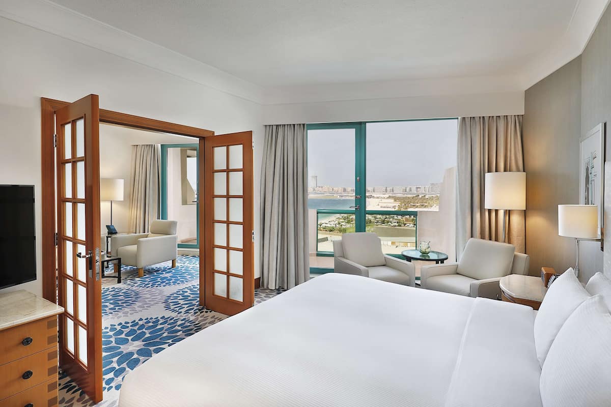 A hotel room with a large bed, two armchairs, a desk with a lamp, and a view of a cityscape through a large window.