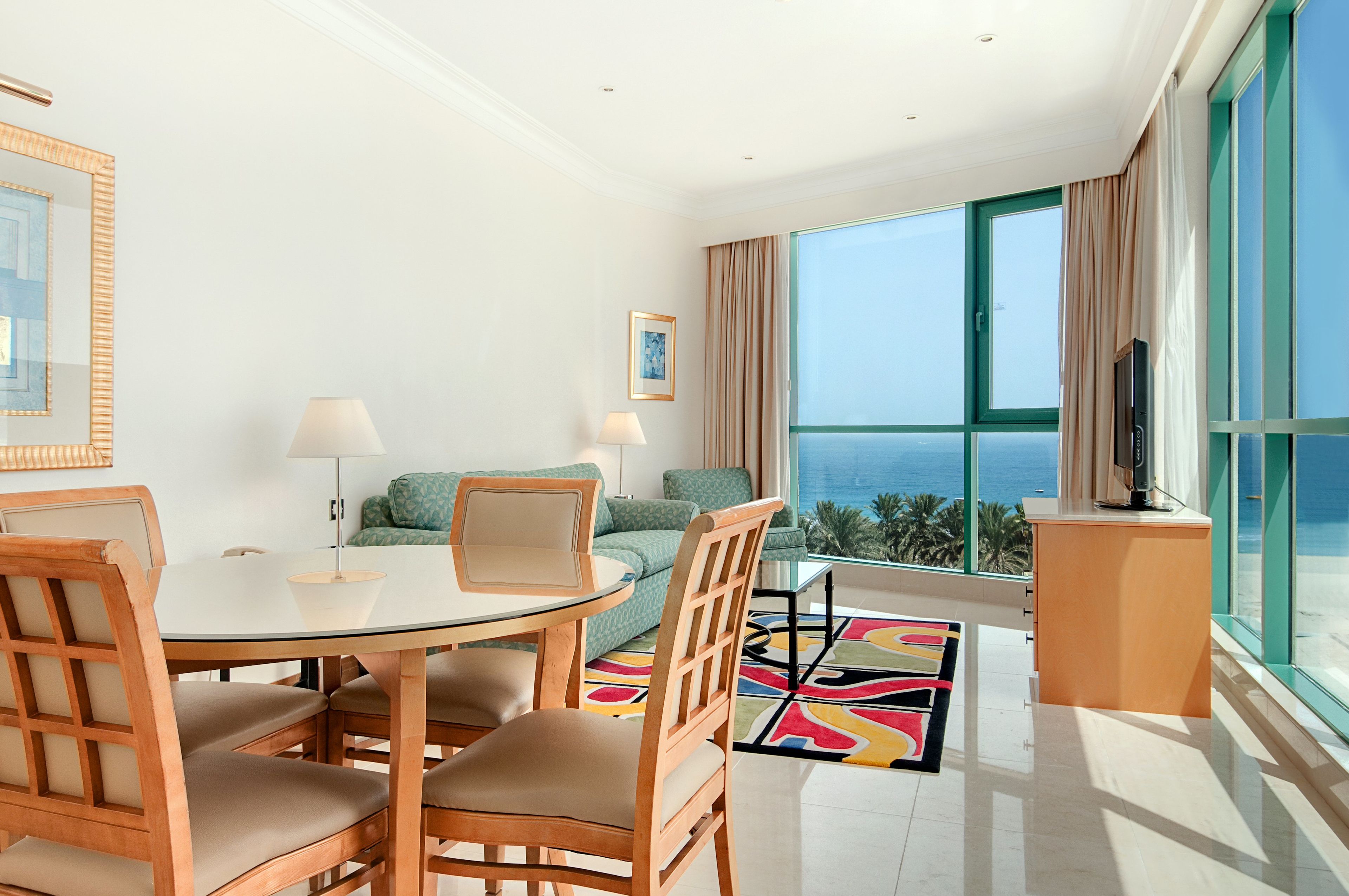 A modern hotel room with a glass table, wooden chairs, a sofa, a TV, and a large window offering a view of the ocean.