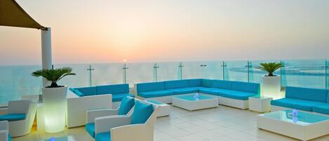 A rooftop lounge with white wicker furniture and blue cushions, overlooking a calm sea at sunset.