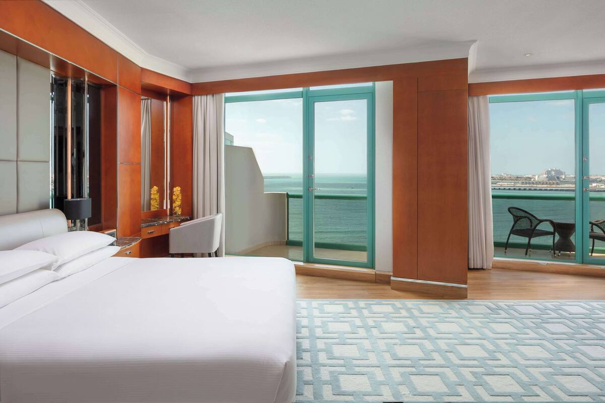 A hotel room with a large bed, a desk with a chair, a large window with a view of the sea, and a patterned rug.