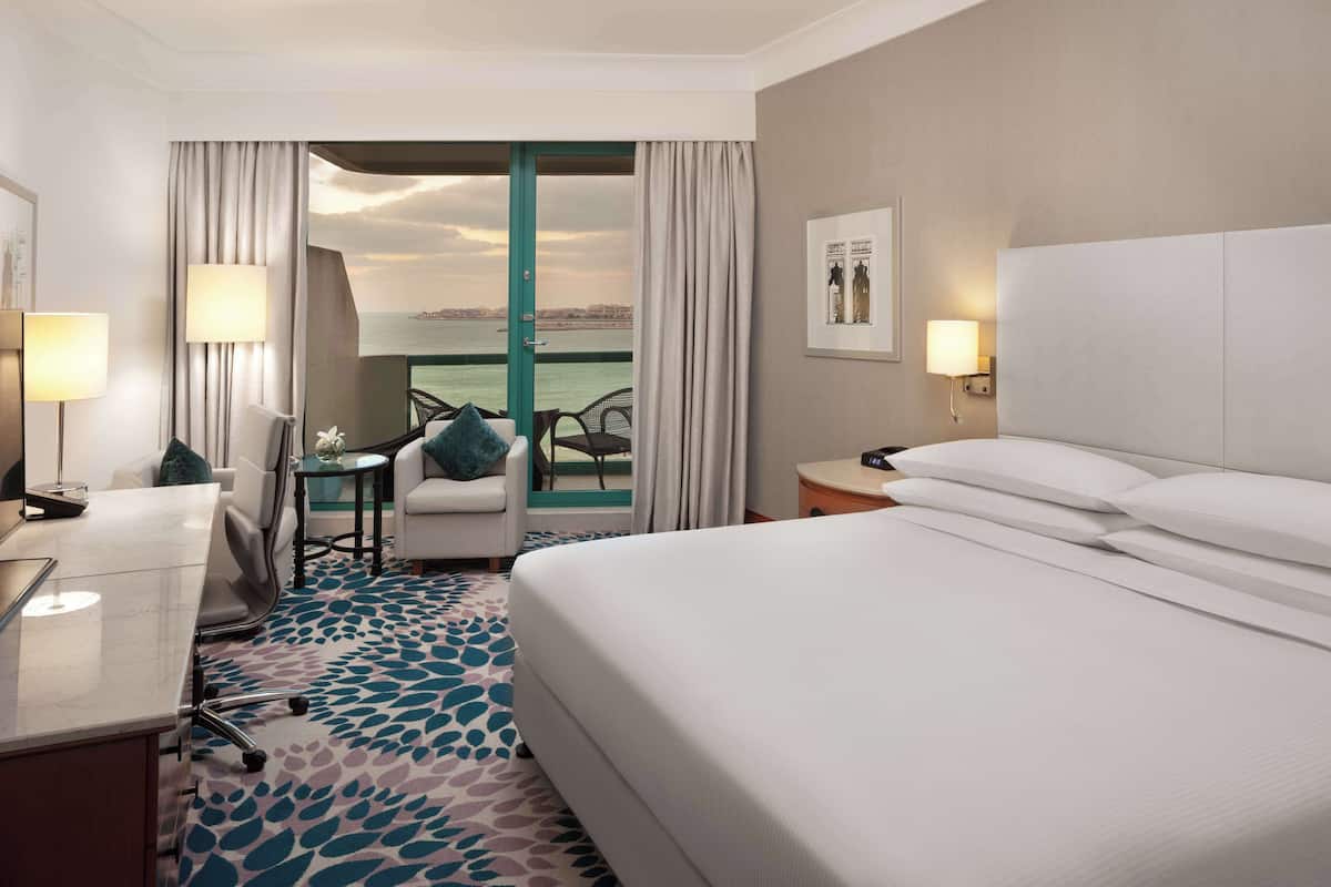 A hotel room with a bed, a desk, a chair, a small table, and a view of the sea.