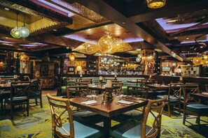 Un restaurante con mesas y sillas de madera, una zona de bar y luces decorativas colgantes.