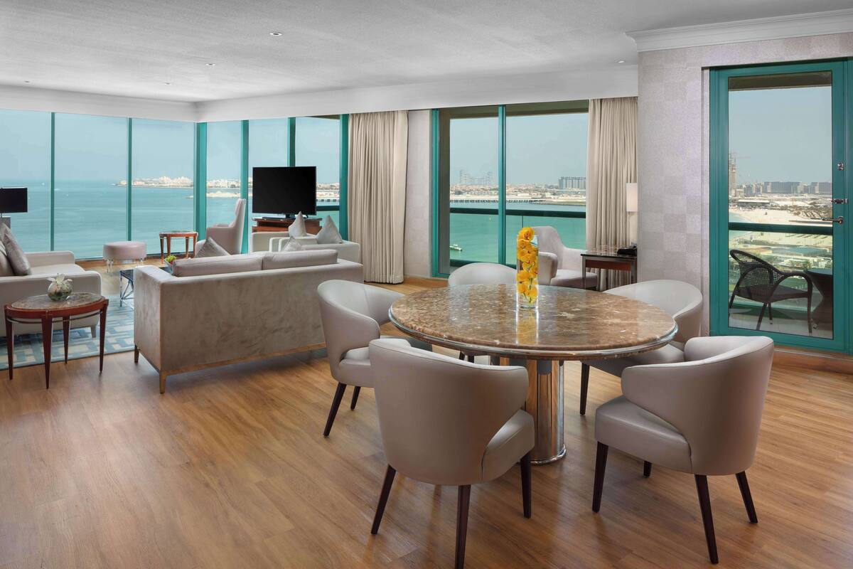 A spacious living room with a large round table and comfortable seating, a flat-screen TV, and a view of the sea through large windows.