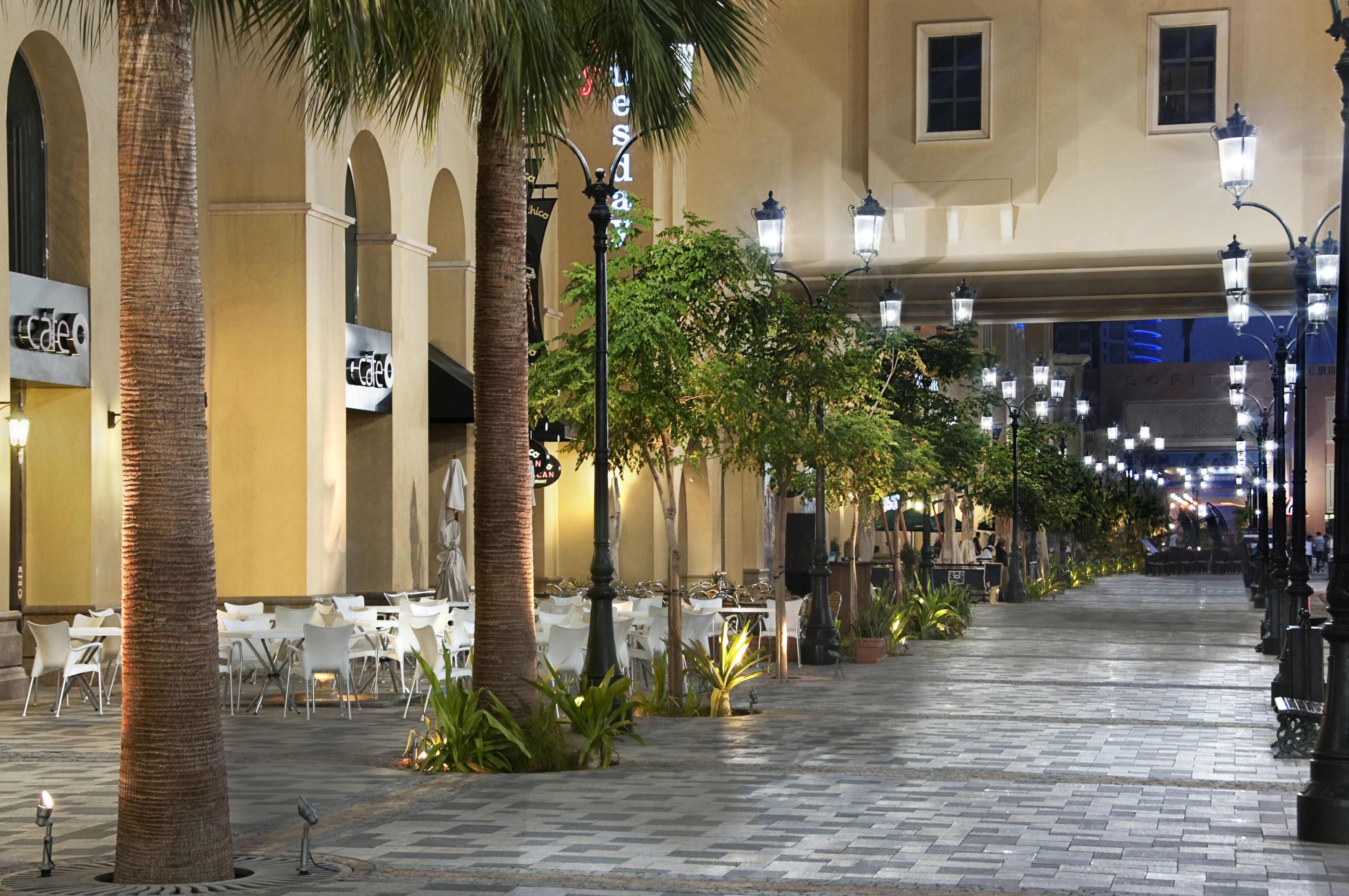 A paved walkway lined with palm trees and white tables, suggesting an outdoor dining area.