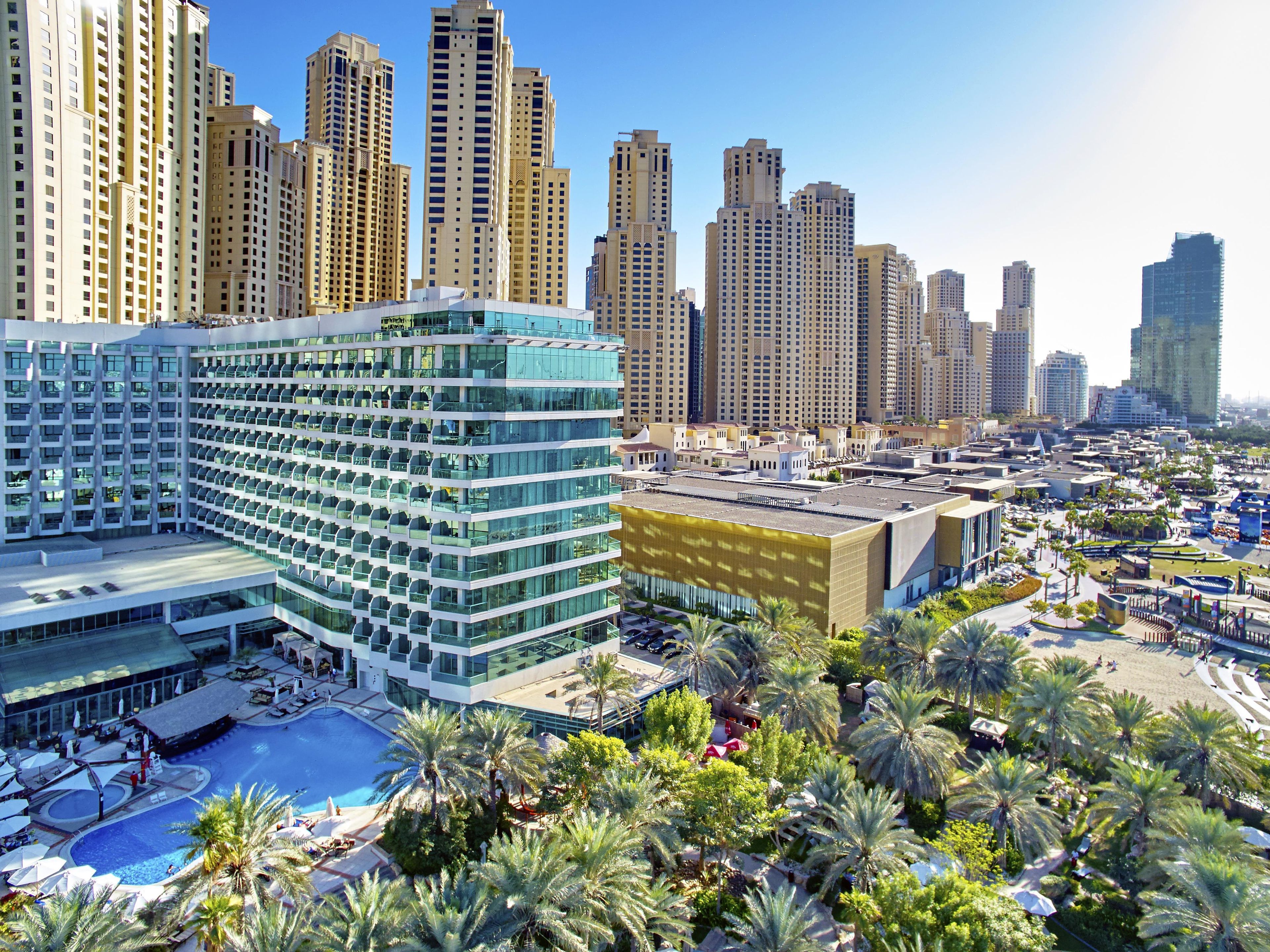 A modern hotel with multiple floors, a rooftop pool, and a landscaped area with palm trees.