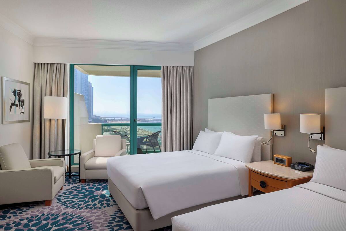 A hotel room with two beds, a desk, a chair, and a view of the city.