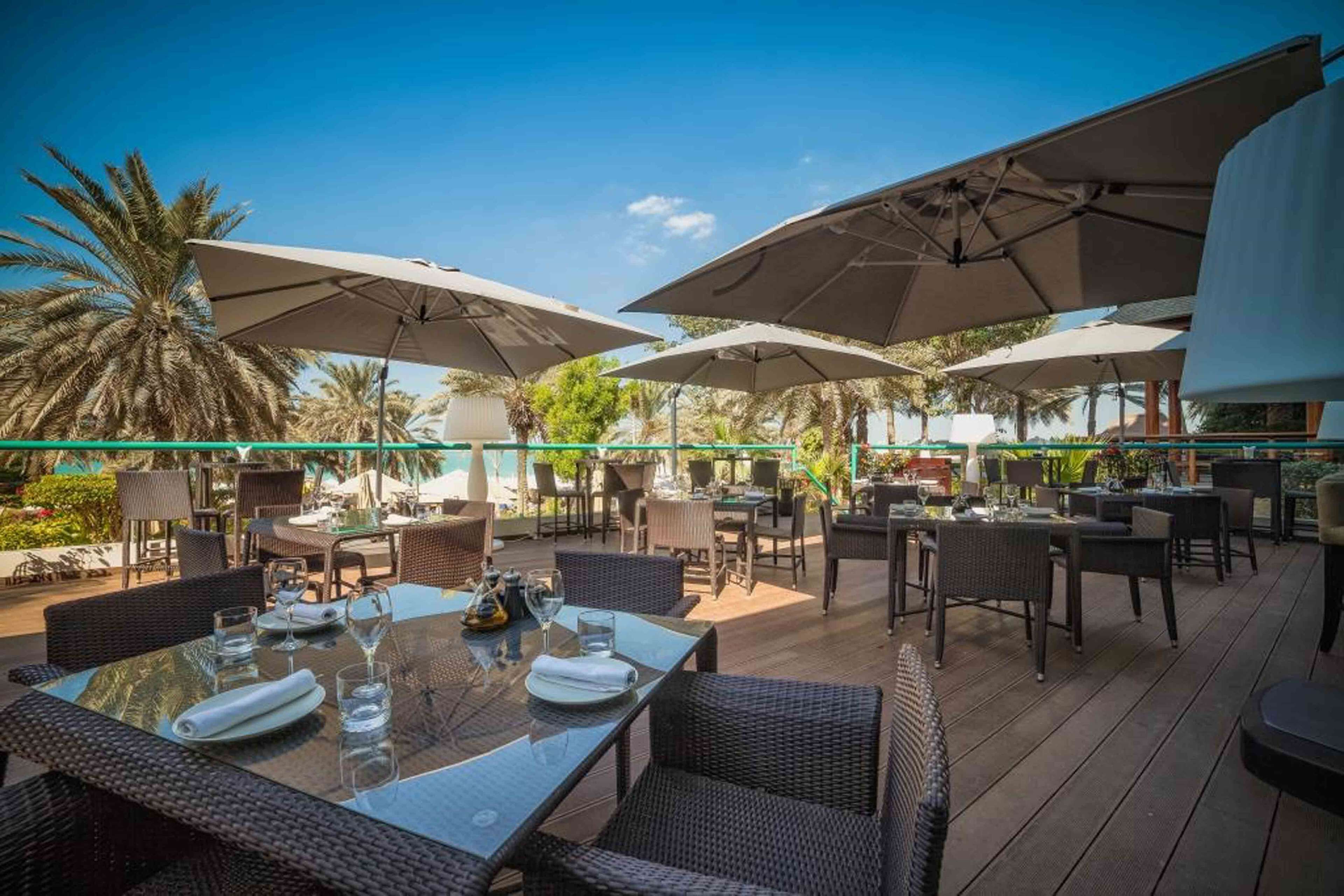 An outdoor dining area with tables and chairs, large umbrellas, and a pool in the background.