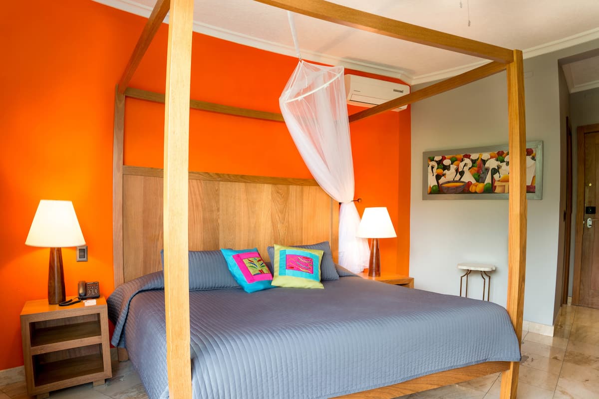 Una cama con dosel, dos mesitas de noche con lámparas, un cuadro en la pared y una puerta al fondo.