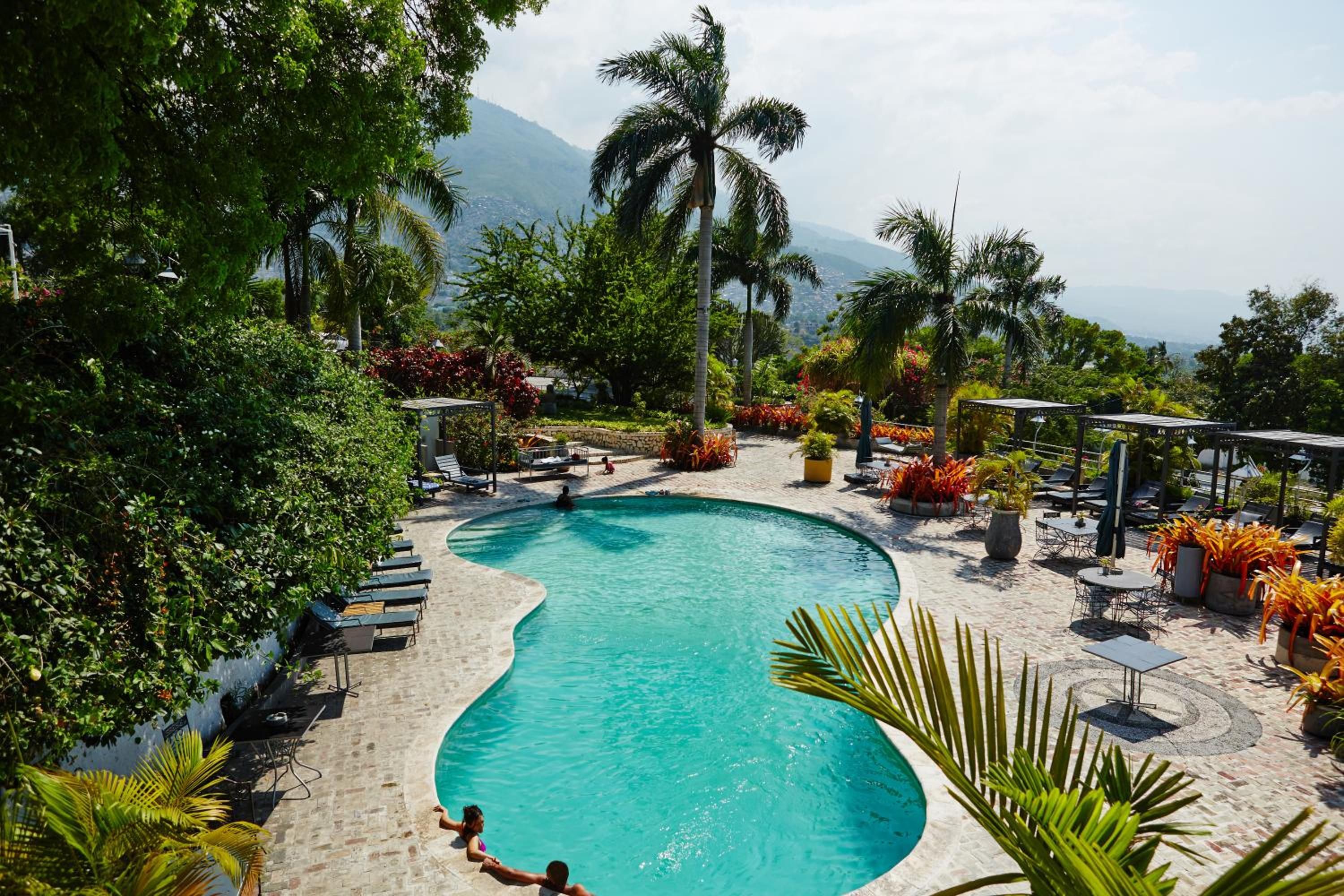 A curved pool with lounge chairs and umbrellas, surrounded by tropical plants and a mountainous backdrop.