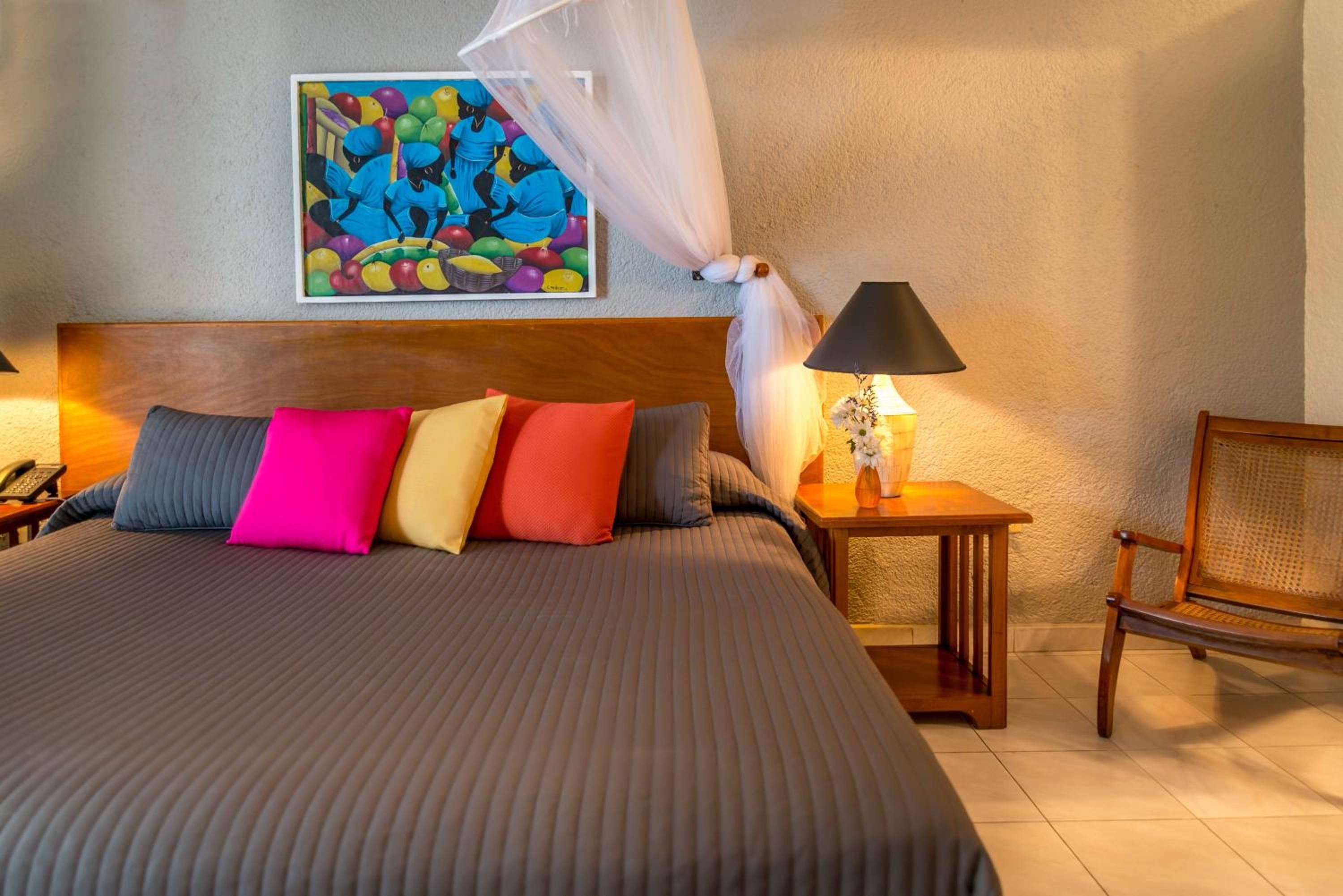 Una cama bien tendida con una colcha a rayas y almohadas de colores, un cuadro en la pared, una lámpara de noche y una silla.