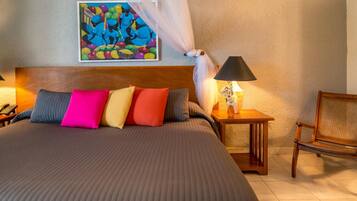 Una cama bien tendida con una colcha a rayas y almohadas de colores, un cuadro en la pared, una lámpara de noche y una silla.