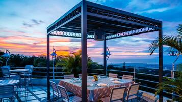 Un espace extérieur aménagé pour les repas, avec une pergola, des tables et des chaises, offrant une vue panoramique sur un magnifique coucher de soleil.