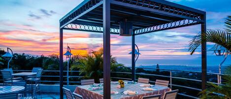 An outdoor dining area with a pergola, tables, and chairs, overlooking a scenic sunset view.