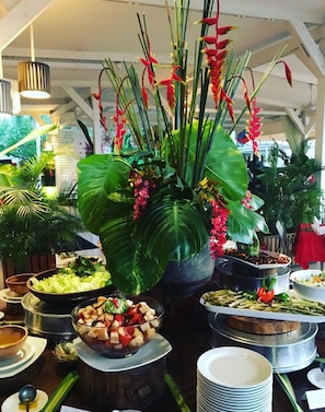 Disposição de buffet com diversos pratos e uma grande planta em vaso com flores vermelhas.