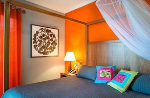 Quarto de hotel com parede laranja, cama com roupa de cama azul, mesa de cabeceira com abajur e um objeto decorativo na parede.