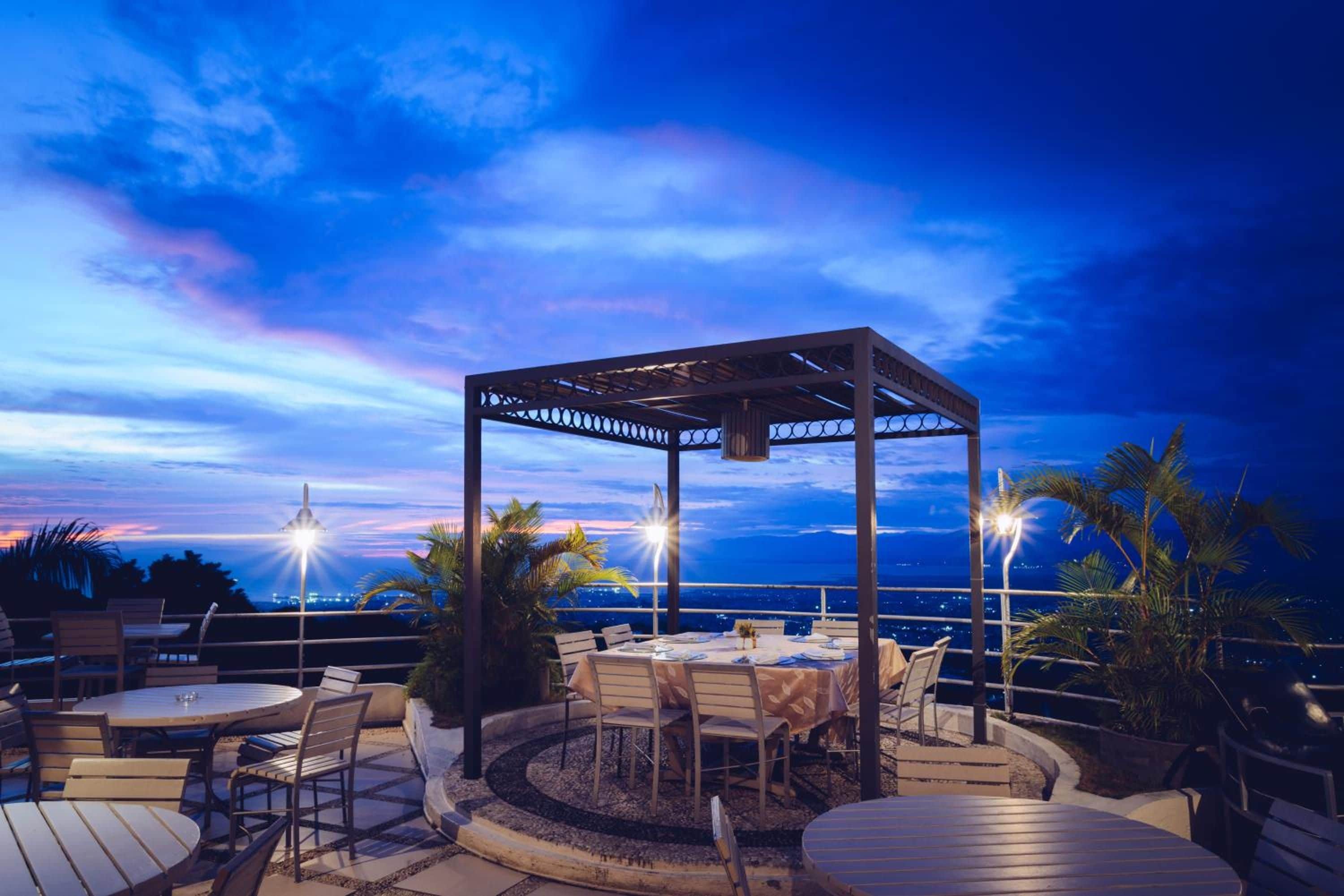 An outdoor patio area with a pergola, round tables, and chairs, overlooking a cityscape at dusk.