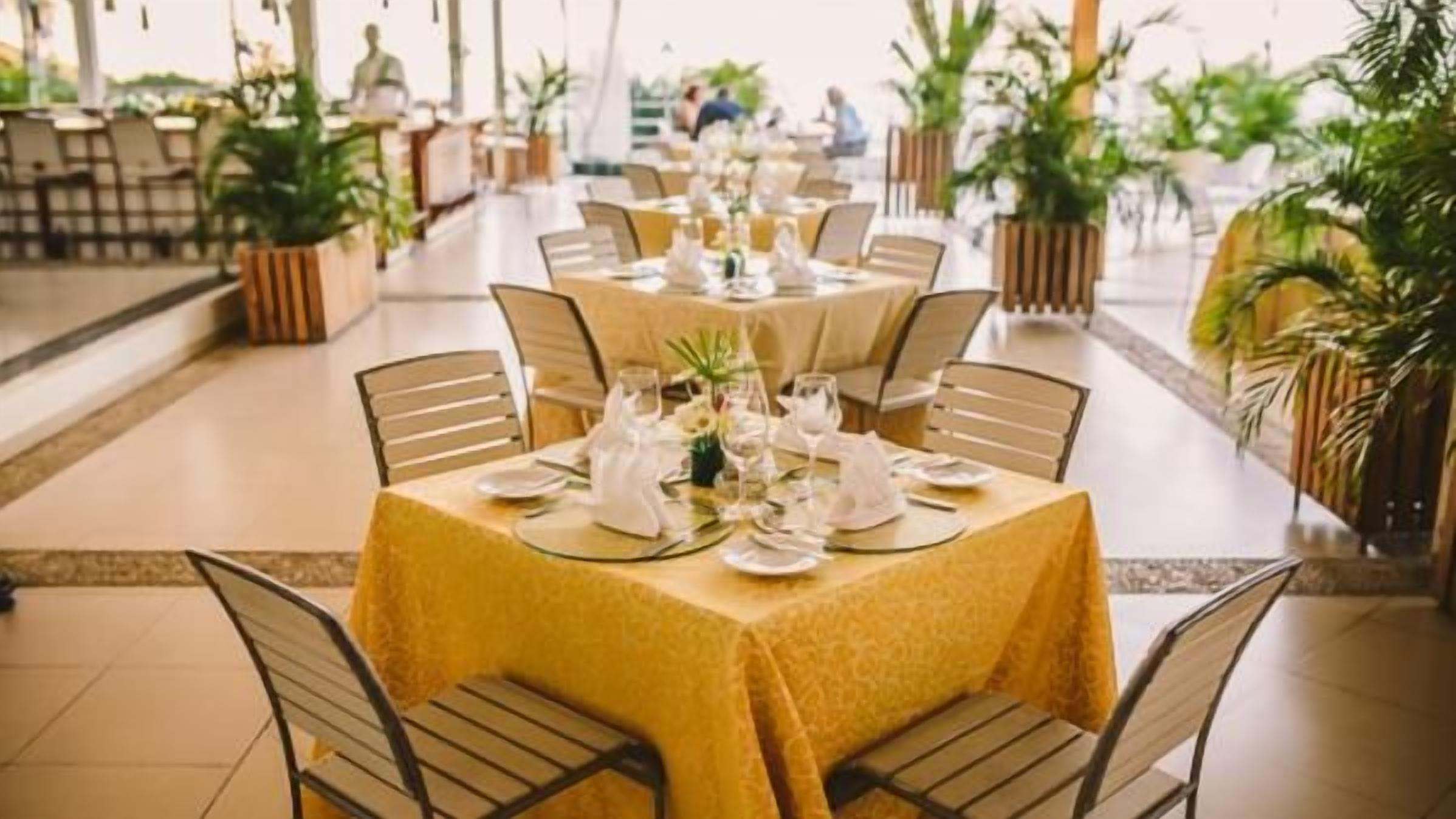 A dining area with round tables set for multiple guests, each table with a yellow tablecloth and white tableware.