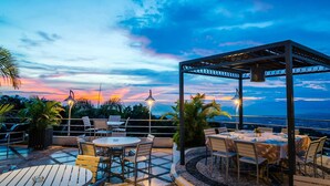 Un espace extérieur aménagé en terrasse, avec des tables et des chaises, une pergola et une vue sur le coucher du soleil.