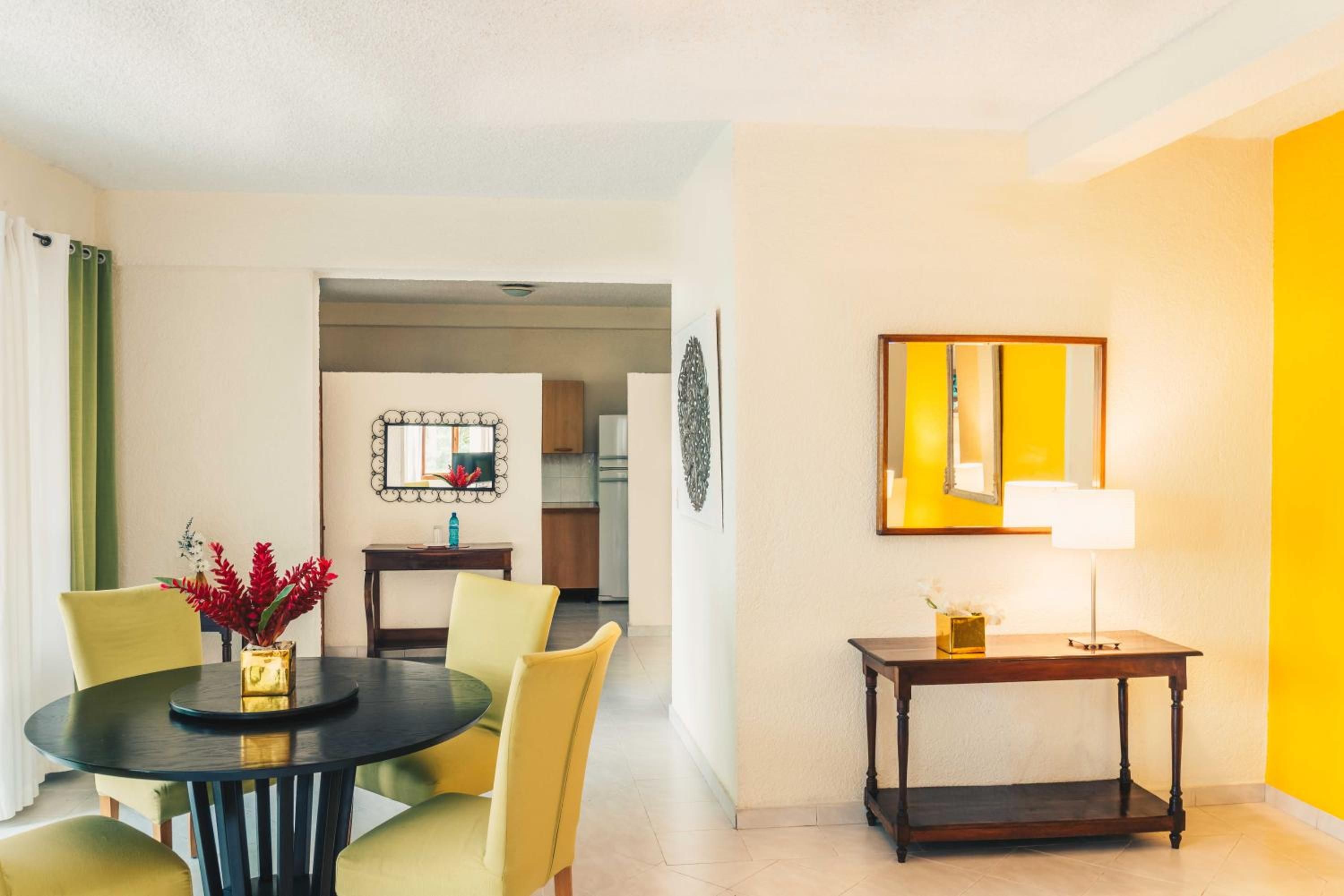 A hotel room with a dining table, chairs, a small table with a lamp, and a mirror on the wall.