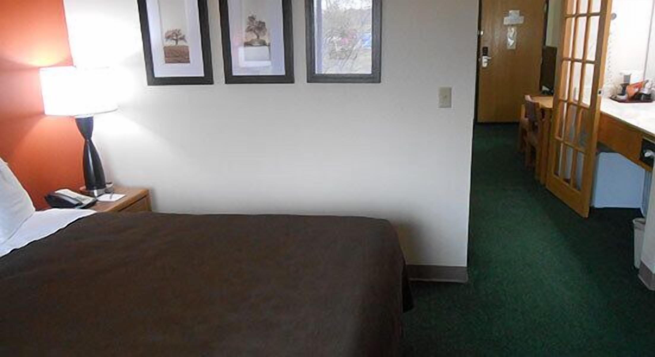 A hotel room with a bed, a desk, a chair, a lamp, and framed pictures on the wall.