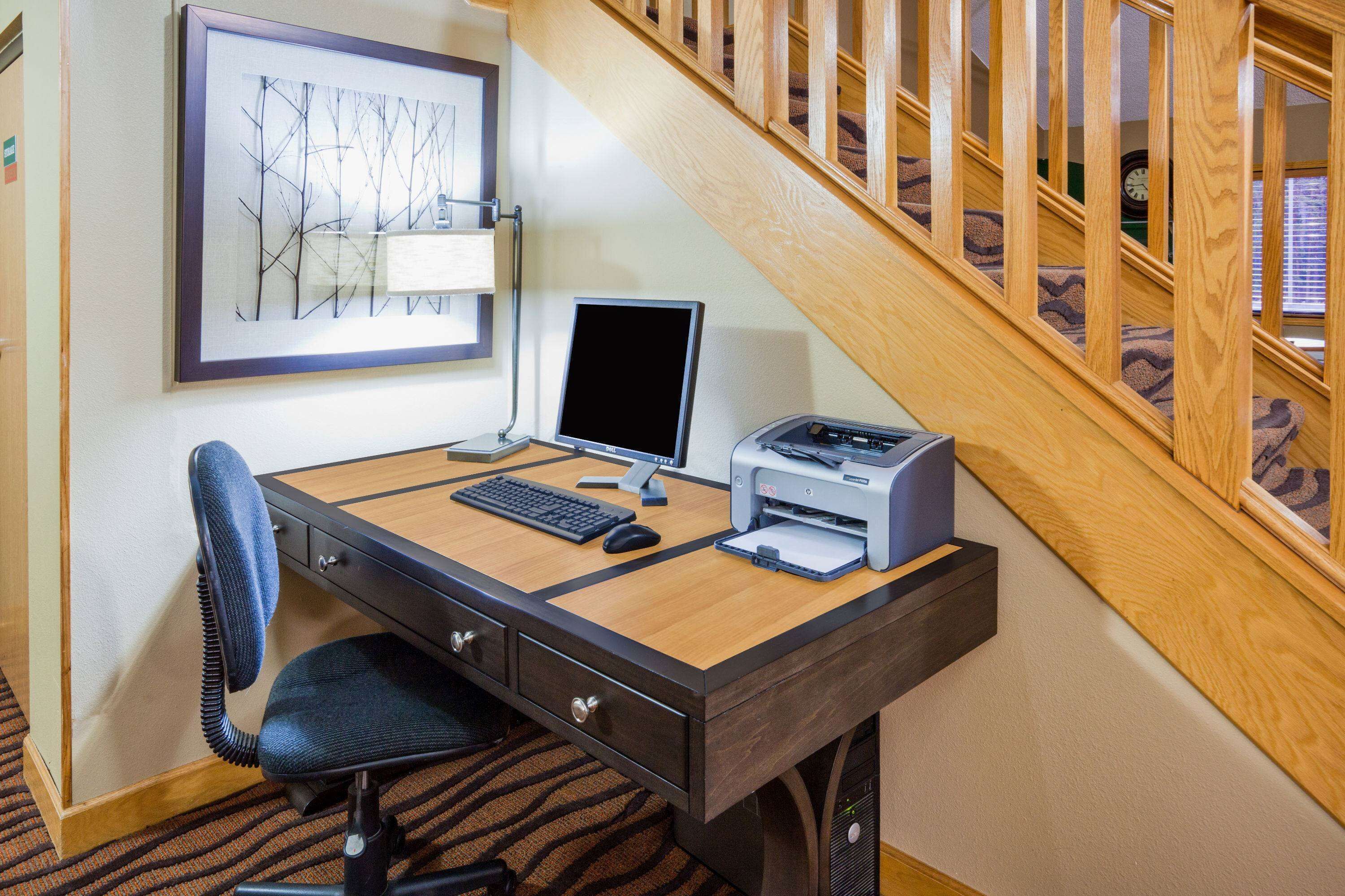 Une chambre d’hôtel avec un bureau en bois, un ordinateur, une imprimante, une chaise et un tableau encadré au mur.