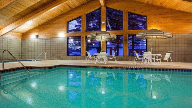 An indoor swimming pool with clear blue water, wooden ceiling beams, and large windows offering views of trees. - AmericInn by Wyndham Mora (Mora)