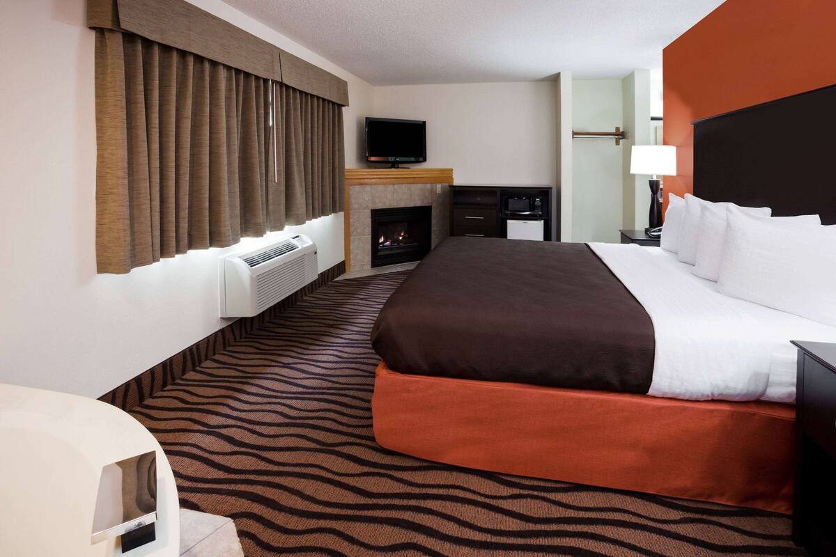 A hotel room with a large bed, a flat-screen TV mounted above a fireplace, a desk with a lamp, and a window with curtains.
