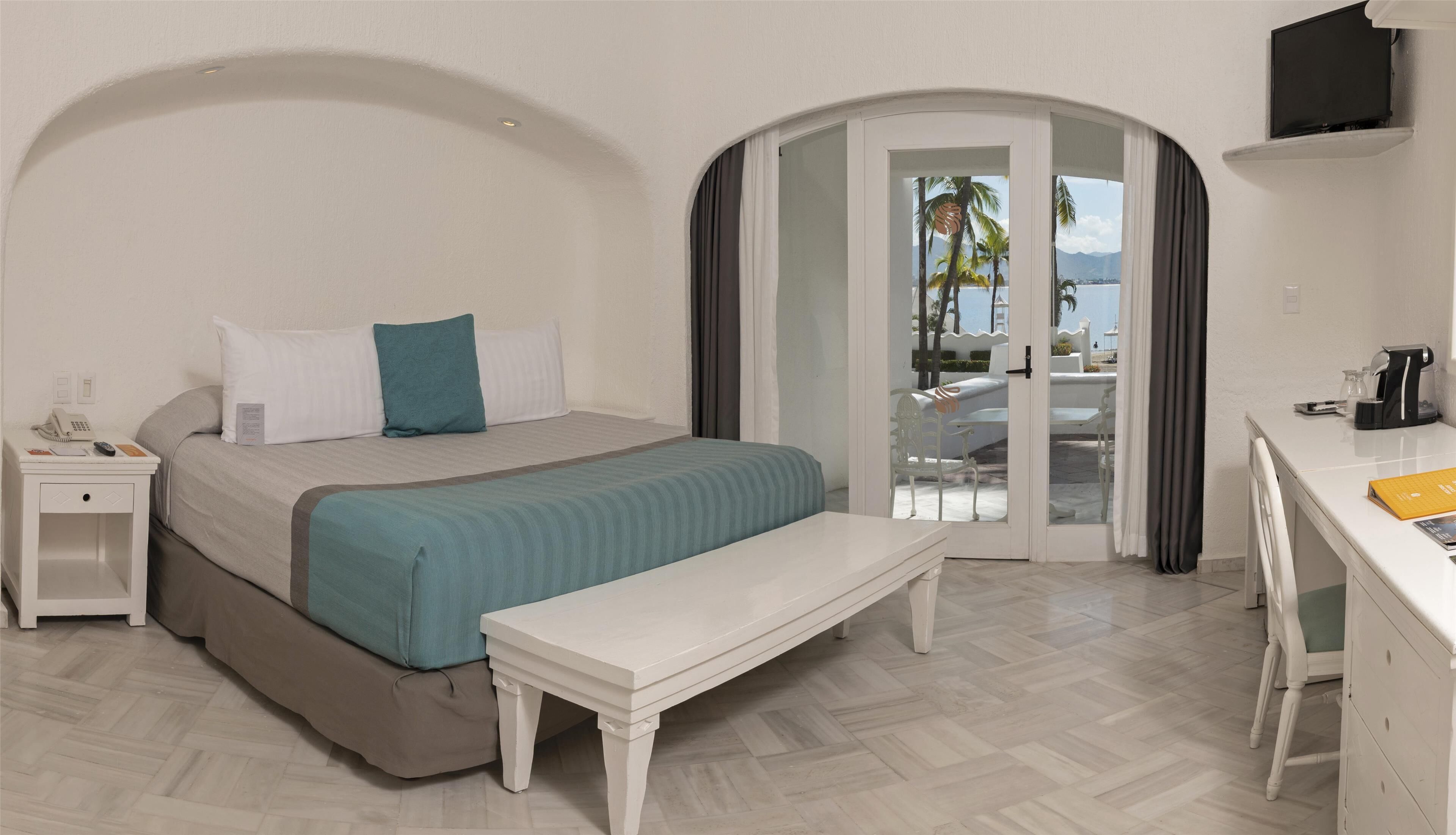 Habitación de hotel con cama, mesita de noche, banco, escritorio y vistas al exterior.