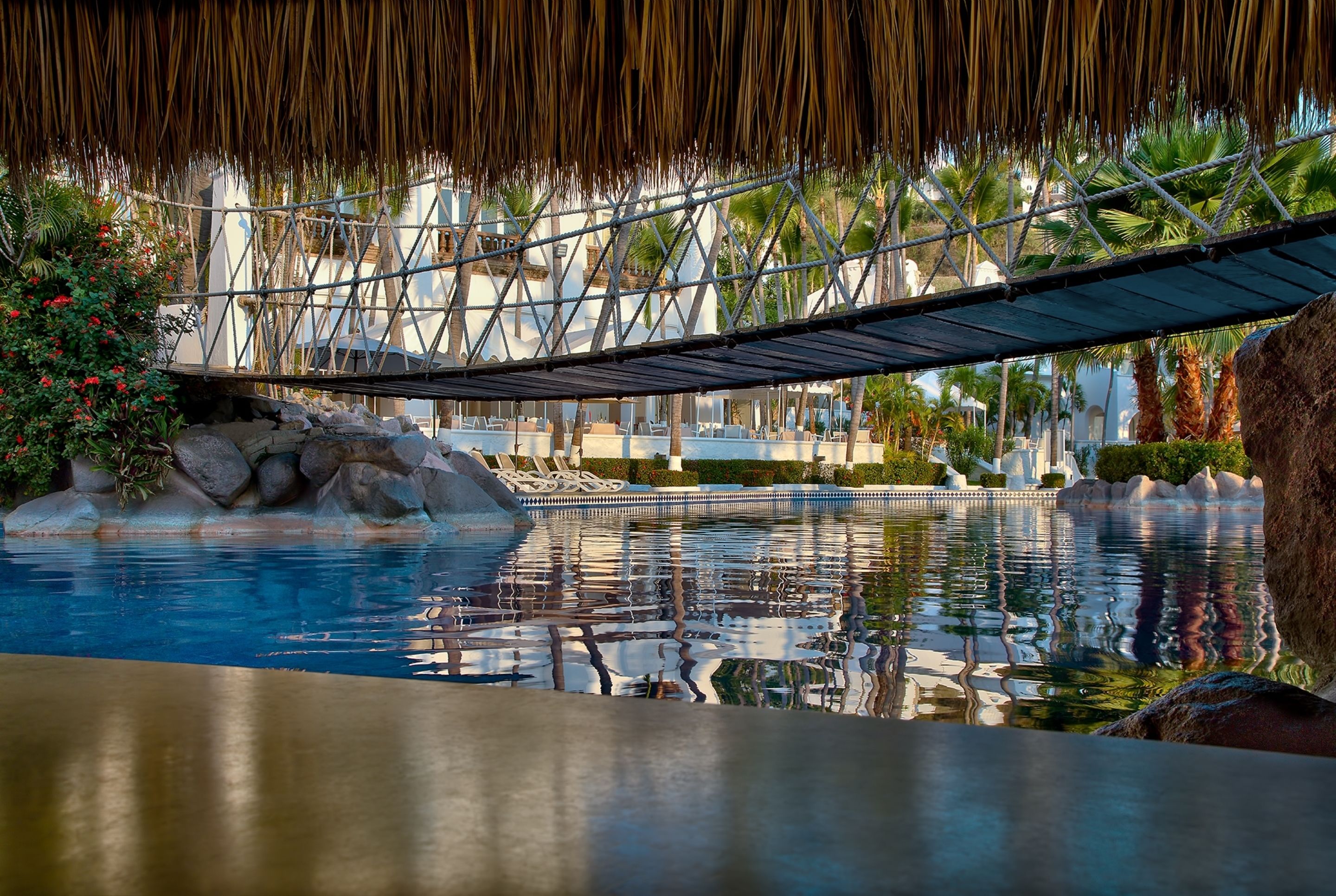 Zona de piscina con un puente, palmeras y una estructura de techo de paja.