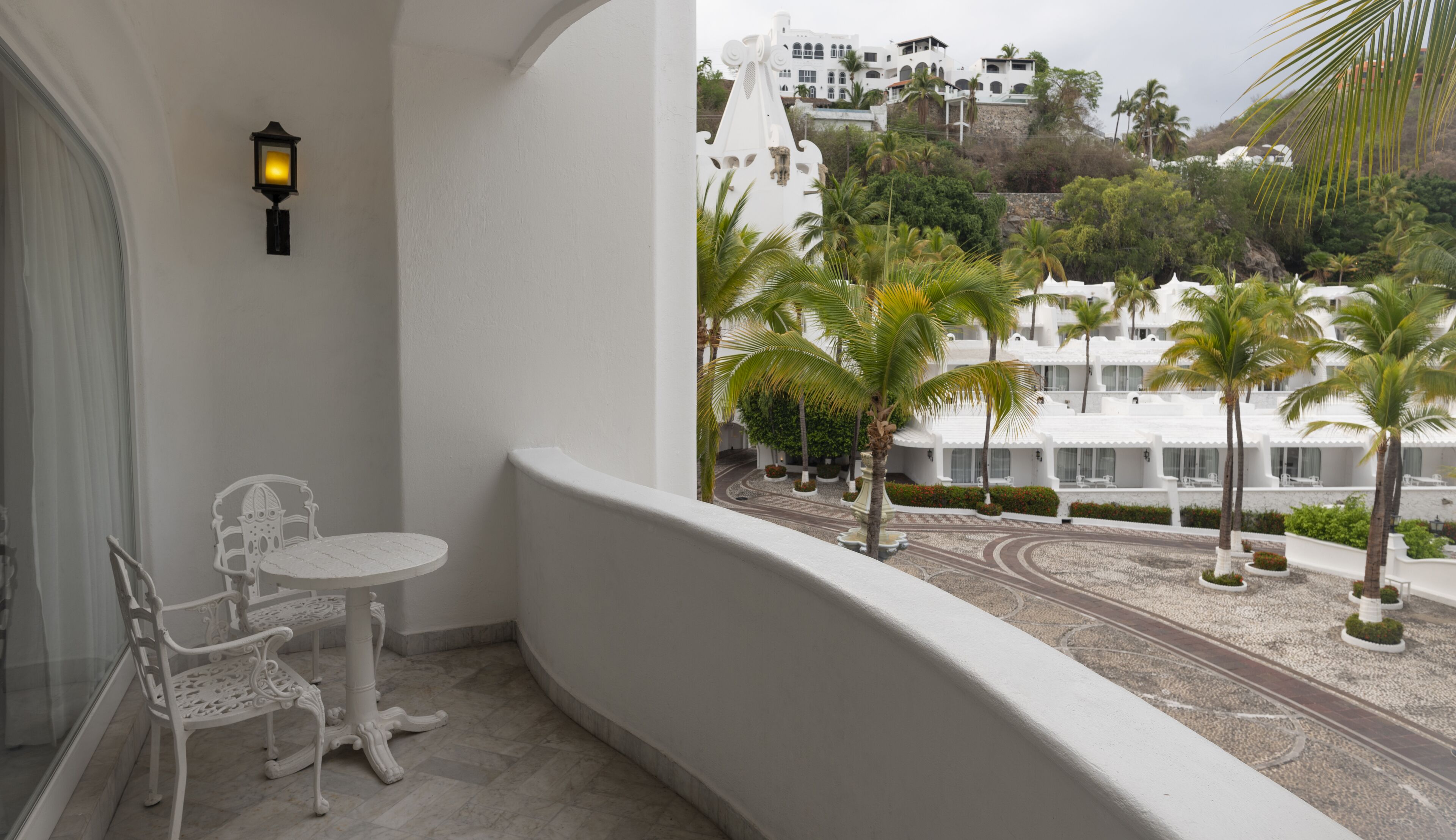 Un balcon avec une chaise et une table blanches, une lampe, et une vue sur un bâtiment blanc, des palmiers et une route sinueuse.