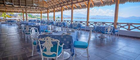 Un restaurante con mesas y sillas, techo de paja y vistas al mar.