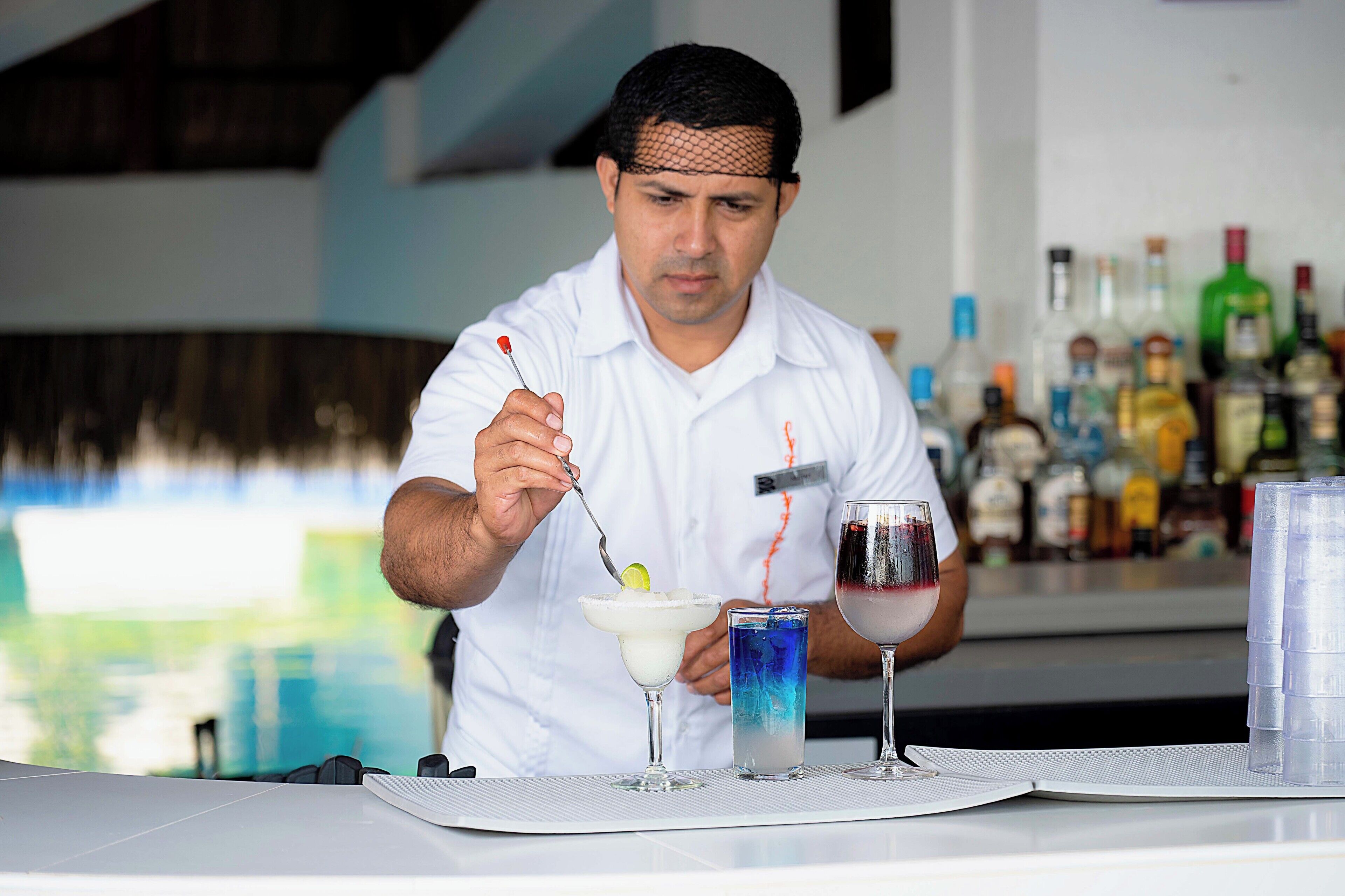 A bartender preparing drinks at a bar with a pool view in the background.