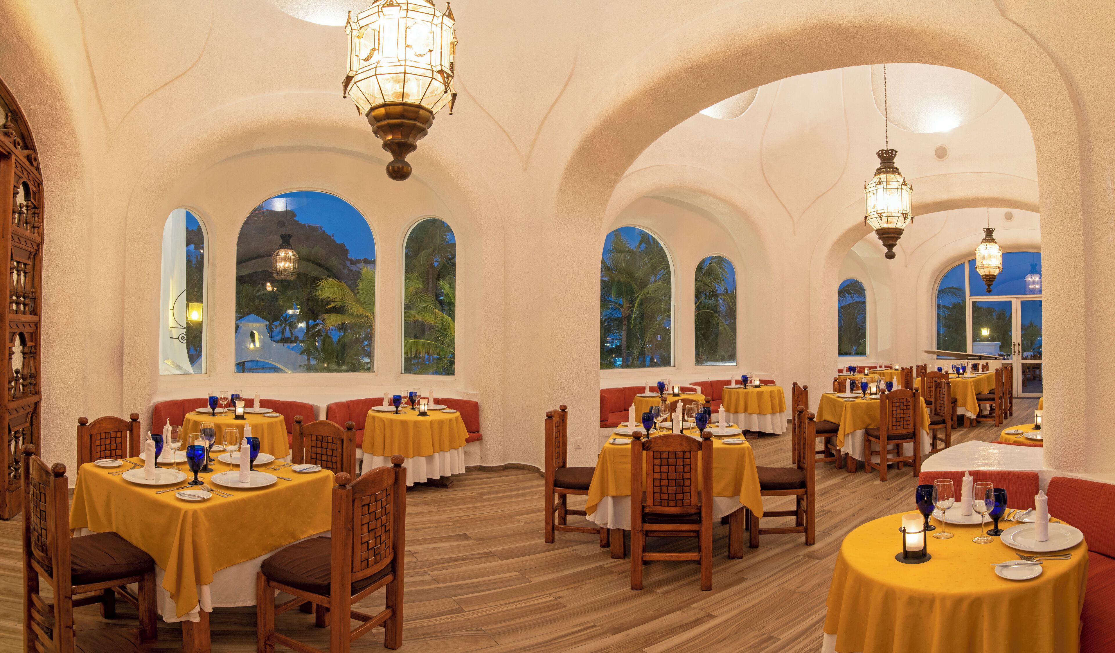 A restaurant with multiple tables, each with yellow tablecloths and wooden chairs. The room has arched ceilings and large windows with views of palm trees and a sunset.