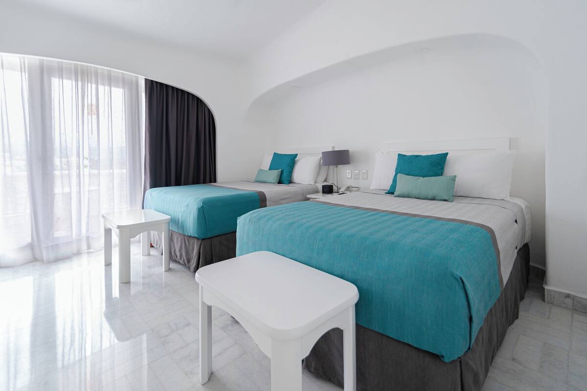 Una habitación de hotel moderna con dos camas, una mesita blanca y un ventanal con cortinas.