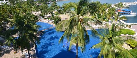 A resort with a large swimming pool surrounded by palm trees, a beach, and a marina with boats.