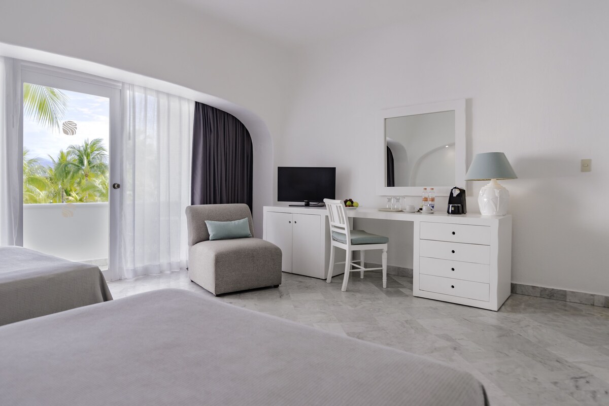 A hotel room with a bed, a desk with a chair, a television, a mirror, and a window with curtains.