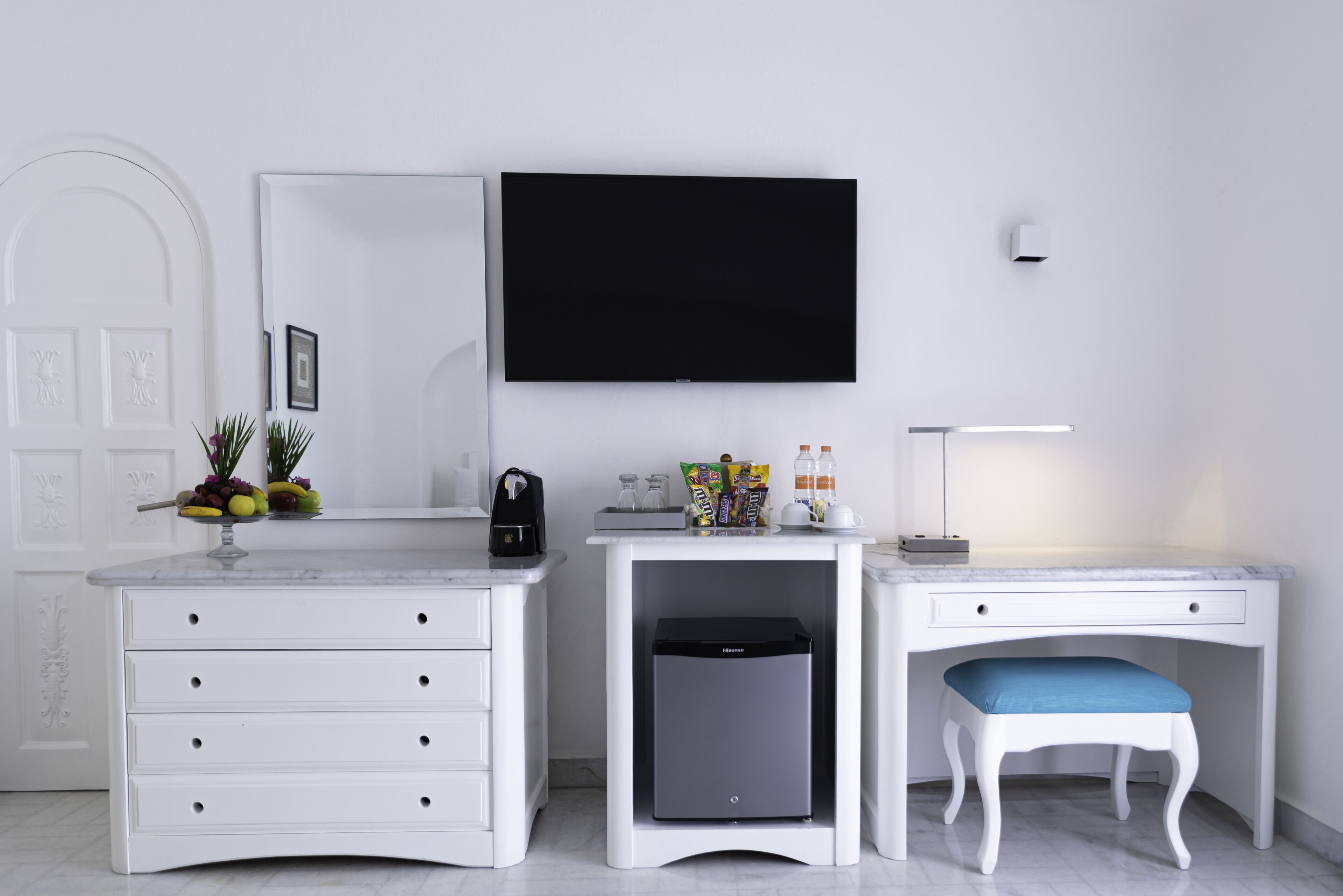 A modern room with a white vanity, a desk with a blue chair, a flat-screen TV, and a small refrigerator.