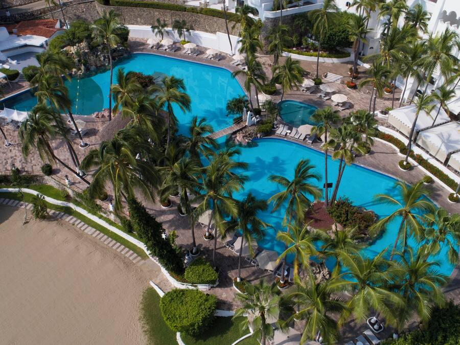 An aerial view of a resort with two large swimming pools surrounded by palm trees and lounge chairs.