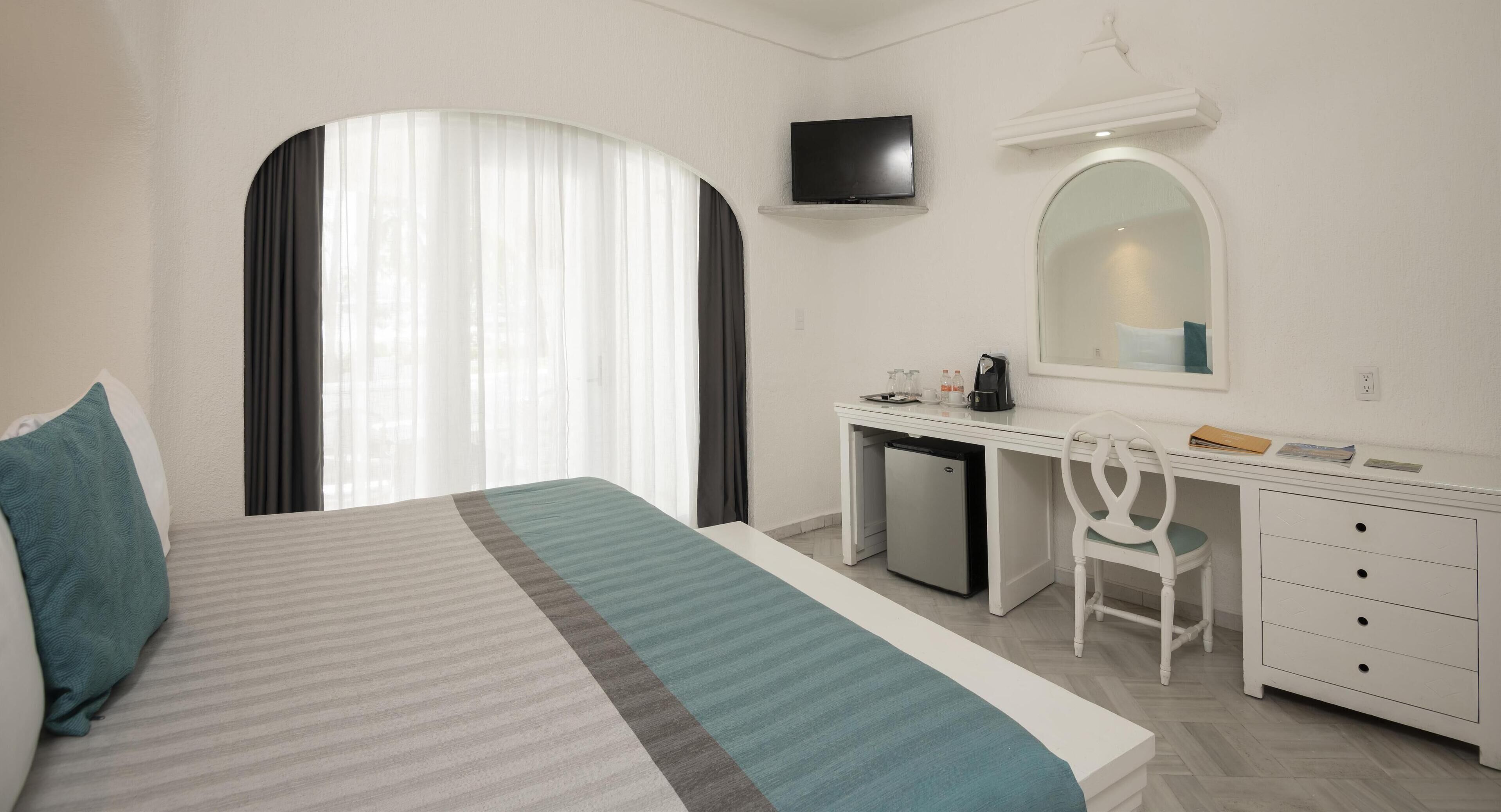 A hotel room with a bed, a desk, a chair, a small refrigerator, a television, and a mirror.