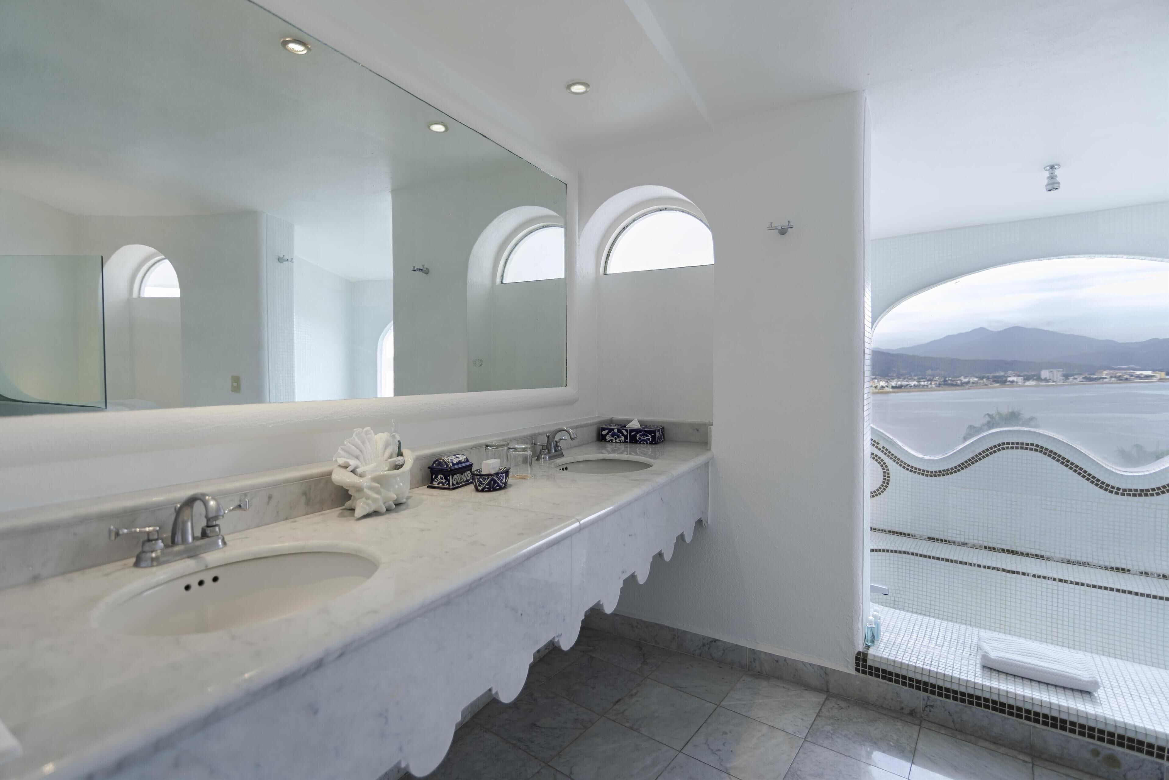Un bagno moderno con un grande specchio, due lavabi e vista sulla città attraverso una finestra ad arco.