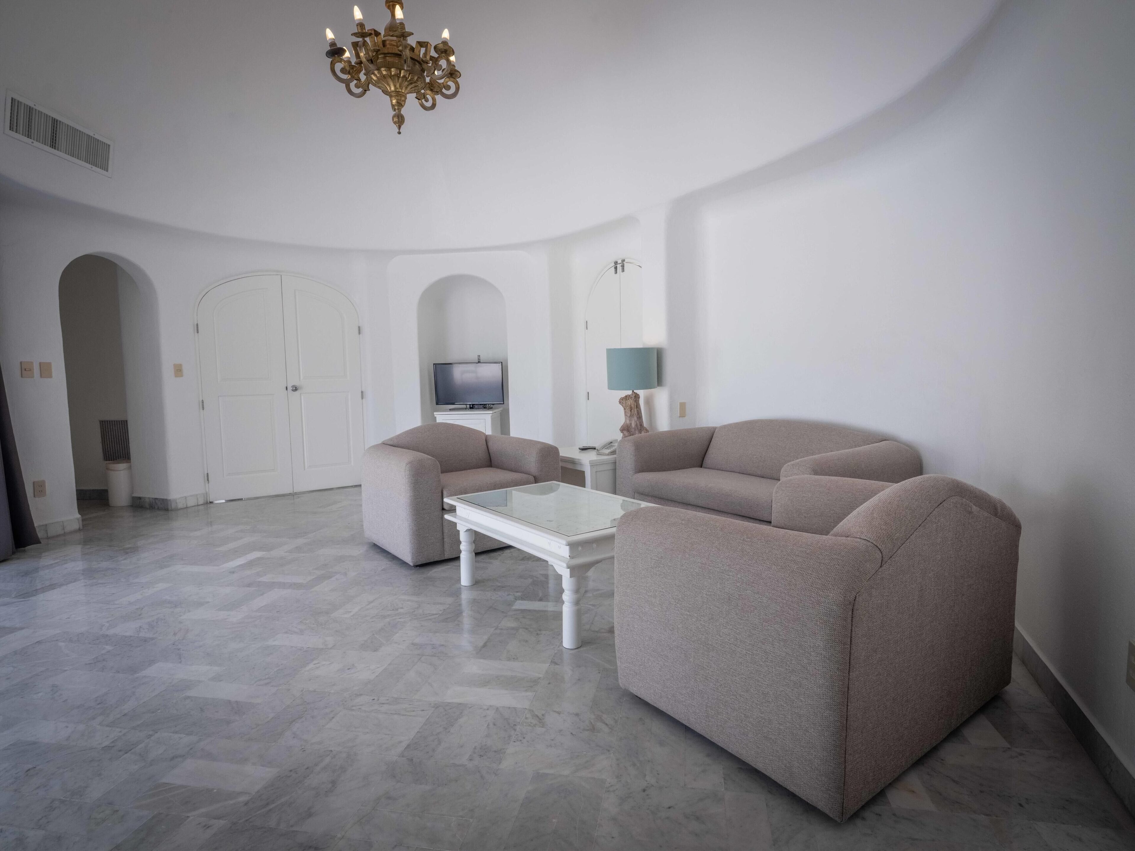 A spacious living room with a modern chandelier, a flat-screen TV, and a sofa set.