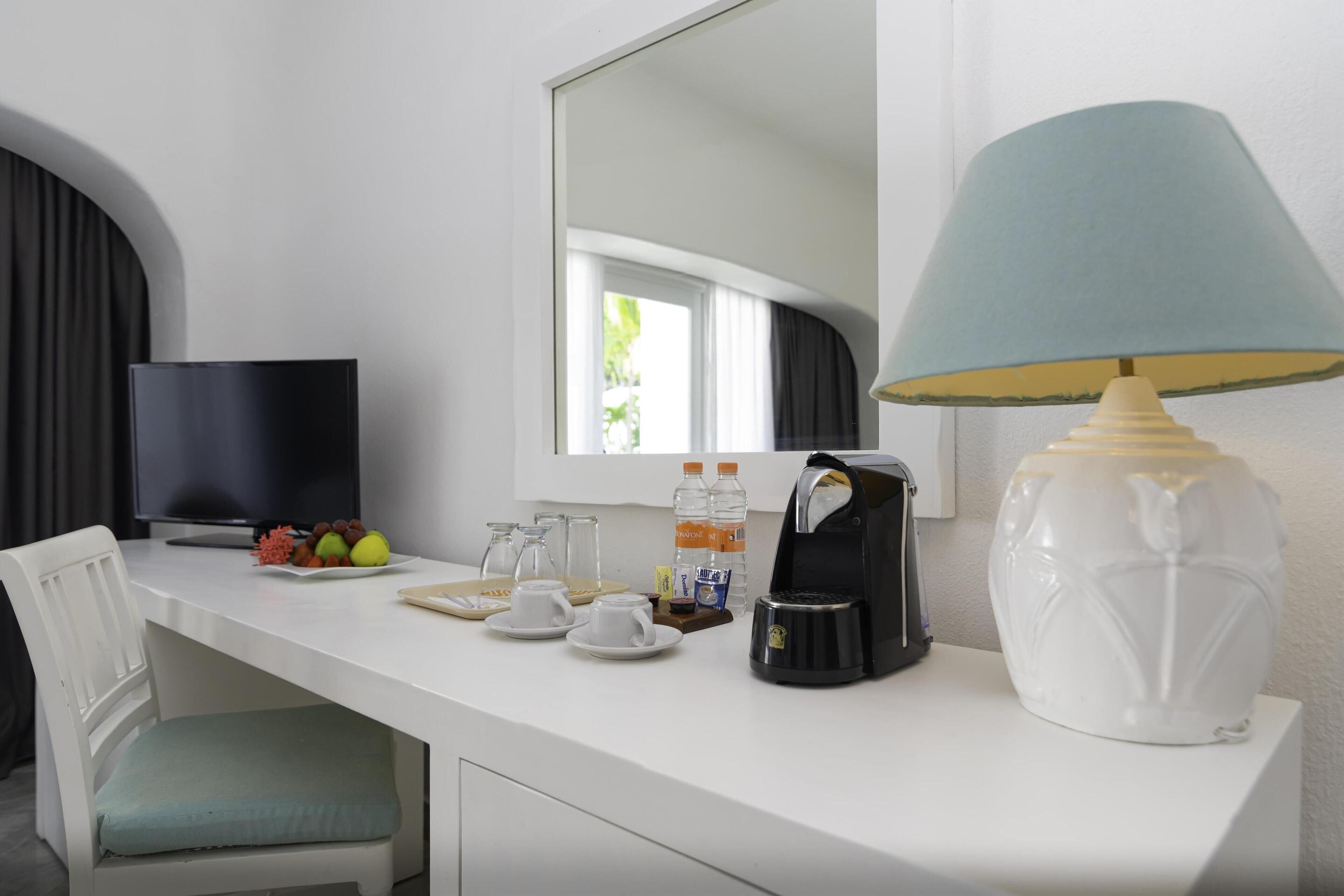 Ein Hotelzimmer mit weißem Schreibtisch, Fernseher, Kaffeemaschine, Lampe und Stuhl.