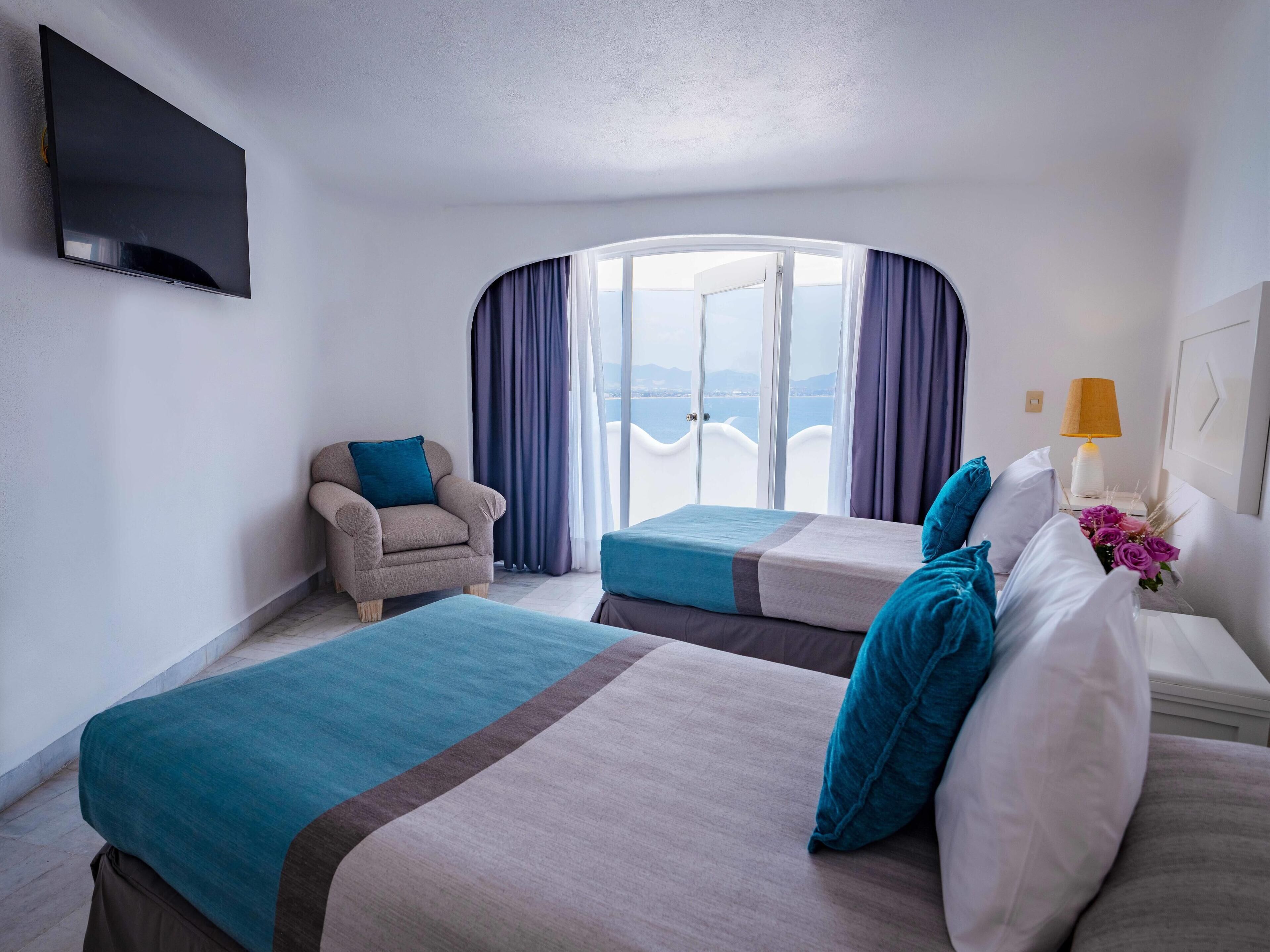 A hotel room with two beds, a chair, a TV, a lamp, and a view of the sea.